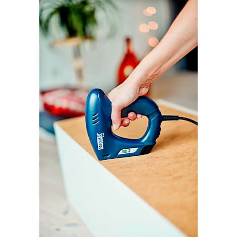 A hand holds a blue stapler on a light brown surface. A white piece of furniture can be seen in the background.