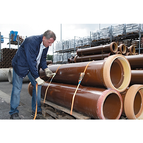 A man in a blue jacket secures brown pipes with straps. In the background, shelves with more pipes.