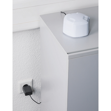 Ein weißer, kompakter Sensor steht auf einem grauen Schrank und ist per Kabel mit einer Steckdose verbunden.
