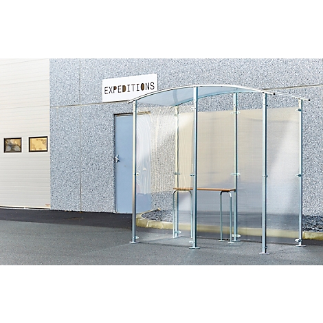 Heller, transparenter Unterstand mit Metallrahmen und Holztisch vor einer Lagerhalle mit Schild "Expeditionen".