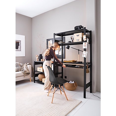 Femme devant une étagère noire avec un bureau. Elle ouvre un tiroir, à côté une chaise. Pièce avec canapé, tapis et décoration.