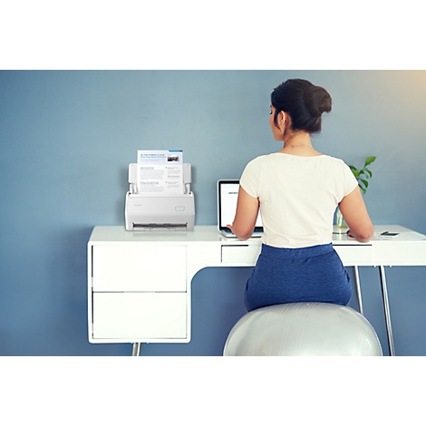 Eine Frau arbeitet an einem Schreibtisch mit Laptop und Dokumentenscanner vor einer blauen Wand.