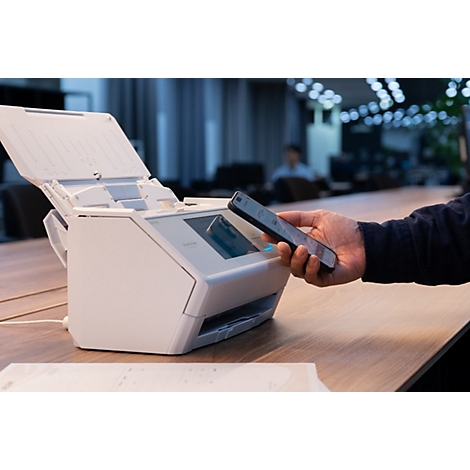 Eine Person hält ein Smartphone über einen weißen Dokumentenscanner auf einem Holztisch in einem Büro.