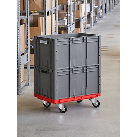 Caja de transporte gris sobre ruedas rojas. Estanterías con cajas en el fondo.