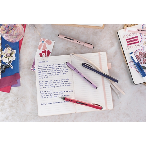 Gros plan sur un carnet, des stylos et des accessoires. Divers stylos reposent sur le carnet ouvert avec du texte.