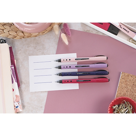 Quatre stylos roses, violets, bleus et rouges sur papier avec des lignes. Articles d'écriture à côté.