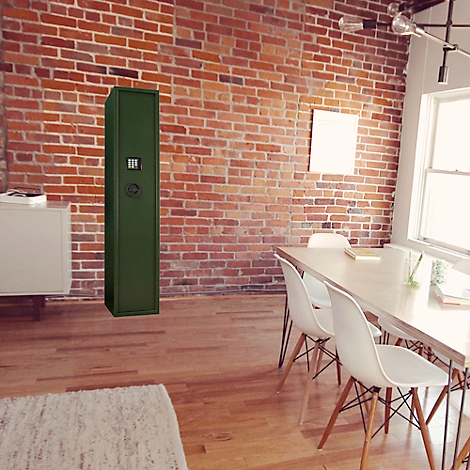 Green tall gun safe in a room with brick wall and wooden floor. A dining table with white chairs stands next to it.