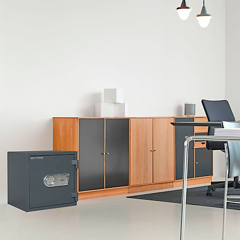 Coffre-fort gris devant un placard dans un bureau lumineux. Au premier plan, une table et une chaise.