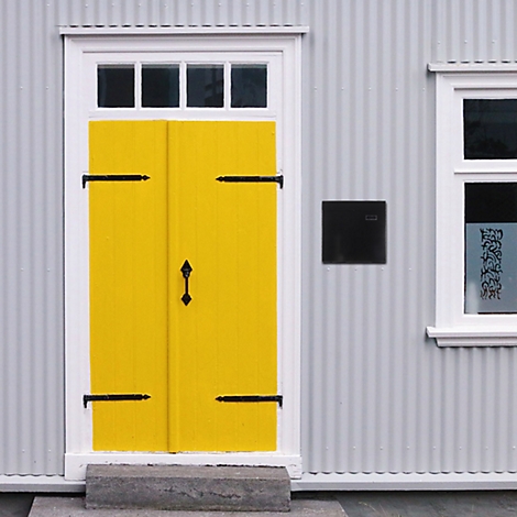 Double porte jaune avec charnières et poignée noires. Mur de maison gris, cadre blanc, petites fenêtres.