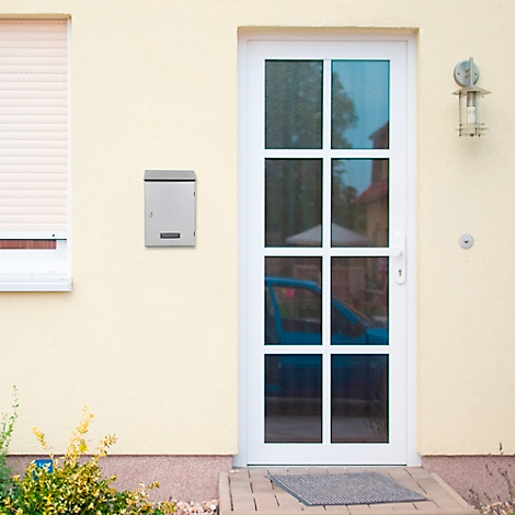 Boîte aux lettres grise sur un mur de maison jaune. Une porte et une fenêtre blanches, ainsi qu'une lampe extérieure sont également visibles.