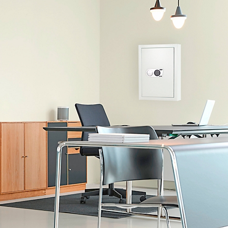 Un coffre-fort blanc est accroché au mur dans un bureau. Un bureau avec une chaise est placé devant le coffre-fort, une commode derrière.