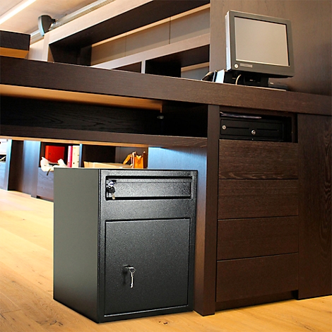 Un coffre-fort noir est placé sous un bureau. Un moniteur et des tiroirs sont également visibles.