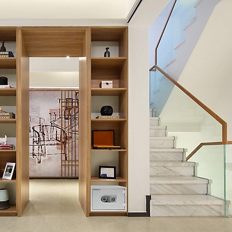 Ein weißer Tresor steht in einem Holzregal, dahinter eine Wendeltreppe mit Glasgeländer.