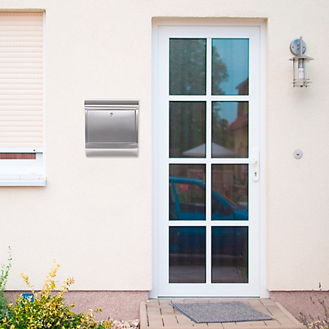 A silver mailbox on a light colored house wall next to a white door. A car can be seen in the background.