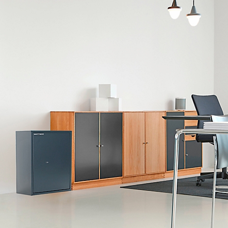 Büro mit Schreibtisch, Schrankwand und Tresor. Helle Wände, grauer Teppich. Deckenlampen.