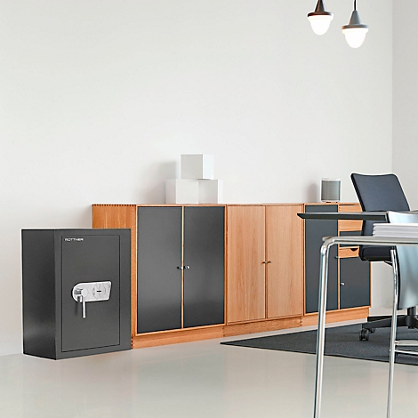 A gray safe stands next to a light wood cabinet in front of a white wall. Two lamps hang from the ceiling.