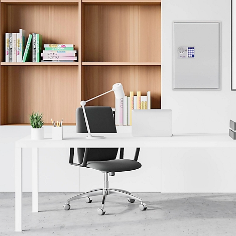 Schreibtisch mit Büchern, Lampe, Bürostuhl und Wandtresor. Bücherregal im Hintergrund. Helle, moderne Büroeinrichtung.