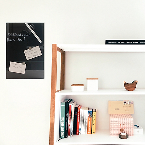 Zwart glazen bord met notities en pen aan een witte muur. Een plank met boeken en decoratie.