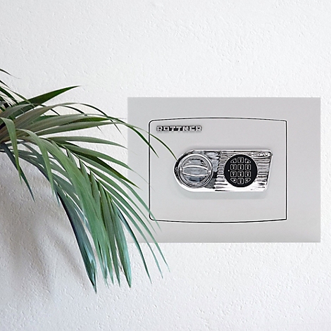 Ein weißer Safe mit Zahlenschloss an der Wand. Daneben grüne Blätter einer Zimmerpflanze.