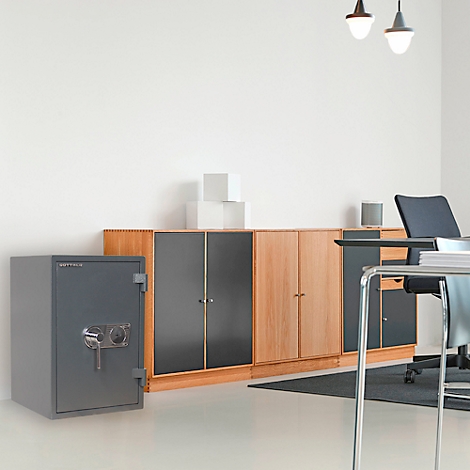 Ein grauer Tresor steht in einem Büro. Dahinter Schränke, Schreibtisch und Stuhl.