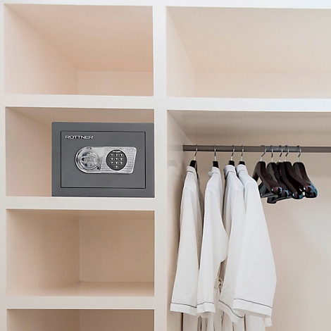 Grauer Tresor mit Zahlencode und Kleiderbügel mit weissen Bademänteln in einem Schrank. Rechts leere Kleiderbügel.
