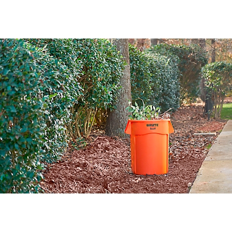 Ein orangefarbener Müllbehälter steht im Garten zwischen Büschen, gefüllt mit Grünzeug, der Name ist sichtbar.