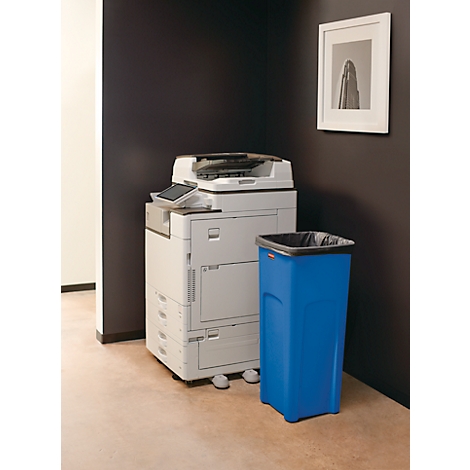 Een witte kopieermachine staat naast een blauwe vuilnisbak in een kantoor. Aan de muur hangt een zwart-wit foto.