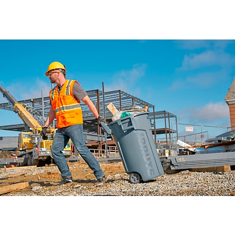 Un ouvrier du bâtiment avec un casque de sécurité tire une poubelle grise sur un chantier.