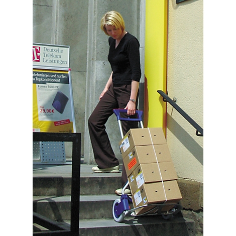 Woman pulls a cart loaded with packages up stairs. In the background an advertisement sign with a telecom logo.