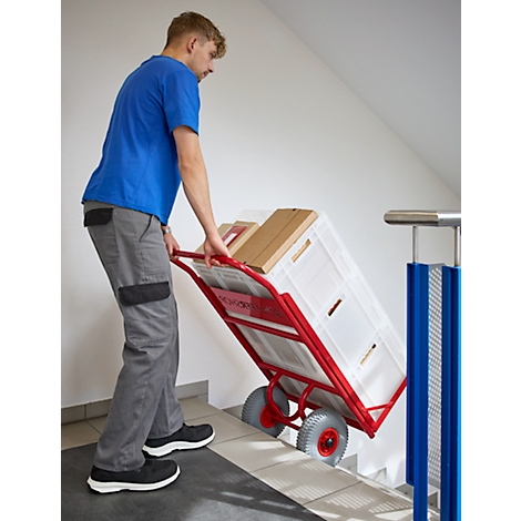 Ein Mann in blauen T-Shirt zieht eine beladene Sackkarre eine Treppe hoch. Auf der Karre befindet sich ein weißer Korb und Kartons.