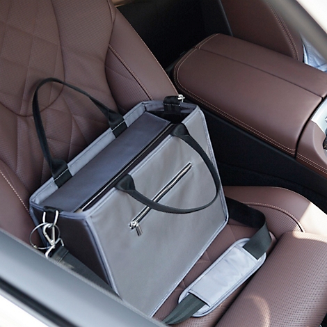 Sac à main gris avec une sangle de transport noire sur un siège de voiture marron. Le sac a une fermeture éclair et une bandoulière supplémentaire.