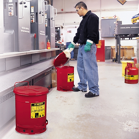 A man is disposing of waste in a red safety can. Another one stands next to it. Industrial environment.