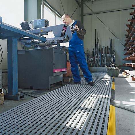 A man in blue overalls operates a saw in a workshop. Gray grid floor, yellow markings.