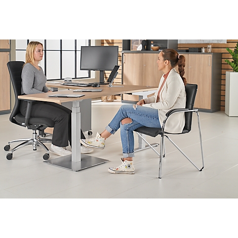 Two women sit at a desk. One wears a grey sweater, the other jeans and a jacket.