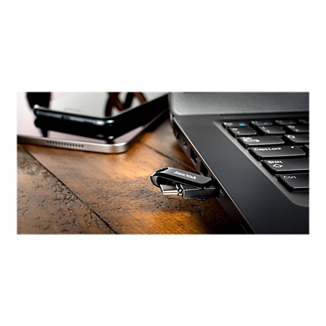 Ein schwarzer SanDisk USB-Stick steckt in einem Laptop, daneben ein Smartphone, beides auf Holz.