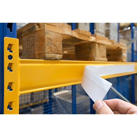 Hand die een zelfklevende witte etiketstrip vasthoudt op een gele schapdrager in een magazijn.
