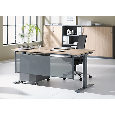 A modern height-adjustable desk. Gray legs and panels. On the light table top are a laptop and office accessories. In the background a window and cabinet.