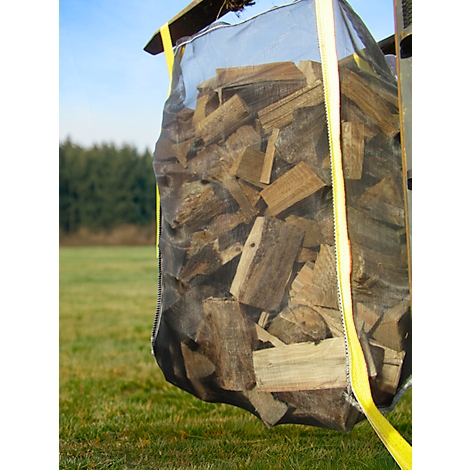 Ein großer, mit Brennholz gefüllter Sack wird von einem Hebezeug gehalten. Auf einer Wiese mit Wald im Hintergrund.