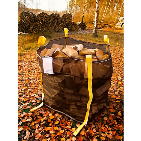 Un grand sac rempli de bois est posé sur des feuilles. Des sangles de transport jaunes sont visibles. En arrière-plan, des tas de bois.