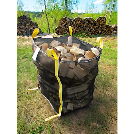 Un grand sac noir rempli de bûches est posé sur une prairie. Des boucles jaunes servent à le transporter.