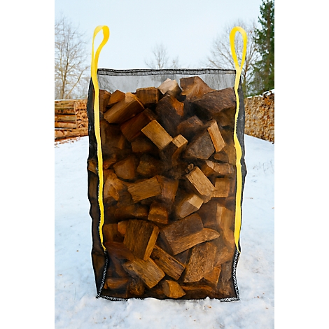 Un grand sac rempli de bois de chauffage. Le sac est noir avec des poignées jaunes. L'arrière-plan montre un paysage enneigé et d'autres piles de bois.