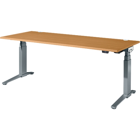 Un bureau réglable en hauteur. Plateau de table brun clair, pieds gris. Les commandes sont situées sur le pied droit.