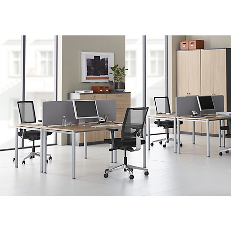 Kantoor met meerdere bureaus, stoelen en computers. Lichte omgeving met kasten en grote ramen.