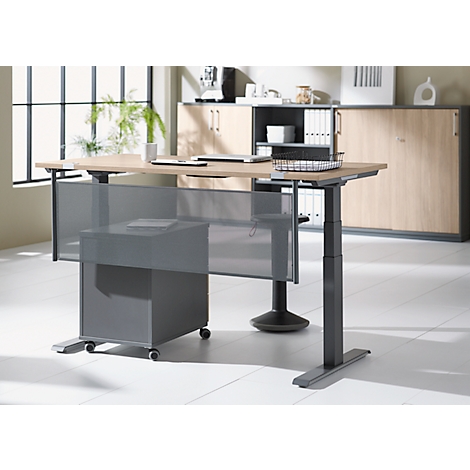 Height-adjustable light brown desk with gray rolling container and privacy screen in a bright office.