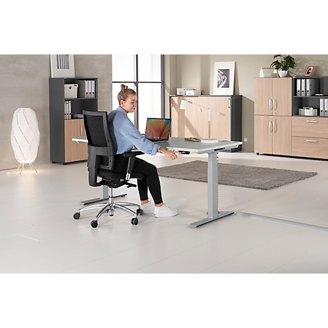 A woman sits at a height-adjustable desk. She operates the desk. Furniture is visible in the background.