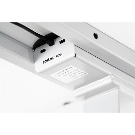Close-up of the underside of a white desk. A white housing with the lettering "SCHÄFER SHOP" and a sticker is shown. A black cable leads into a hole in the table.