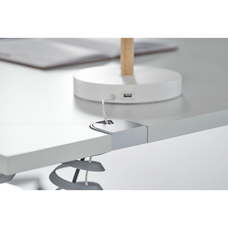 Close-up of a white desk. Part of a table lamp is visible, equipped with a USB port. A book lies on the table.