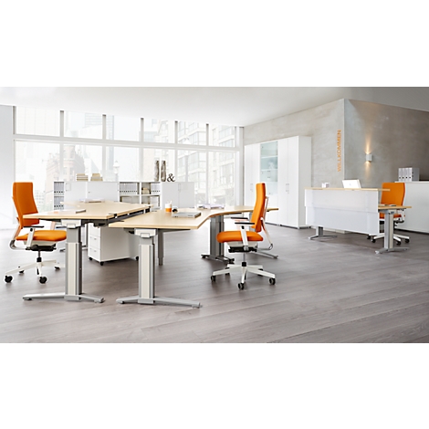 Office space with light tables, orange chairs and wood flooring. Windows and cabinets in the background.