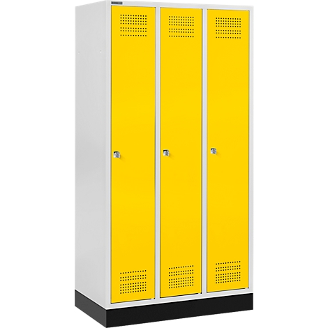 Gele kluisdeuren in drie rijen, naast elkaar. Ze hebben zilveren handgrepen en ventilatiesleuven. De kast is wit en staat op een zwarte basis.