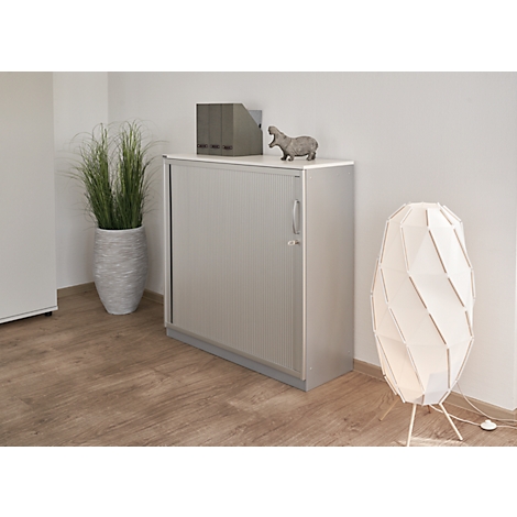 Grauer Schrank mit Lamellentüren, grauer Topf mit Pflanze, Lampe und ein Nilpferd-Figur auf dem Schrank.
